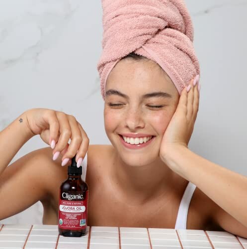 Smiling woman with towel on head holding jojoba oil bottle